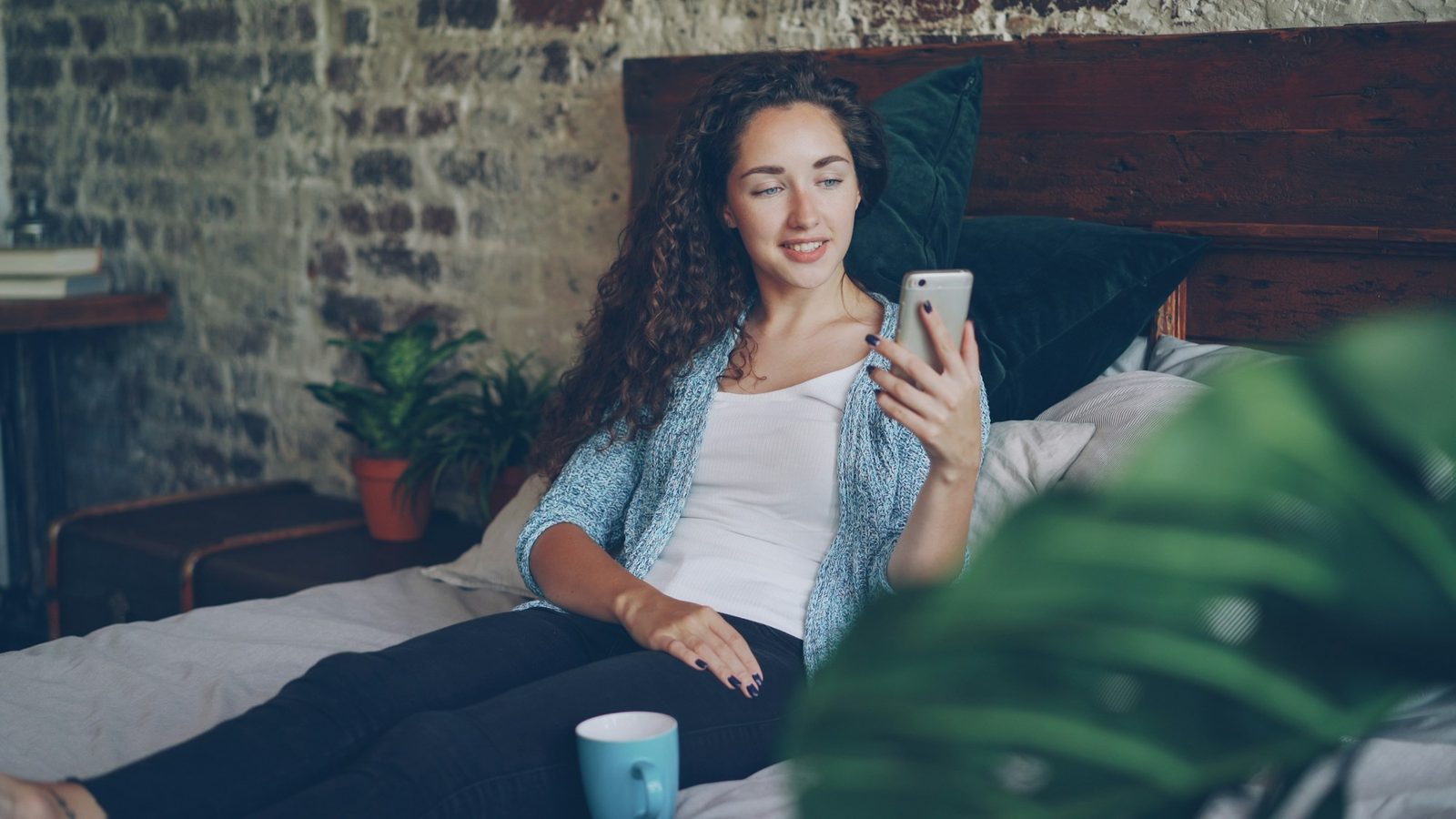 A young woman sitting in bed in the morning, looking at her phone with a coffee beside her