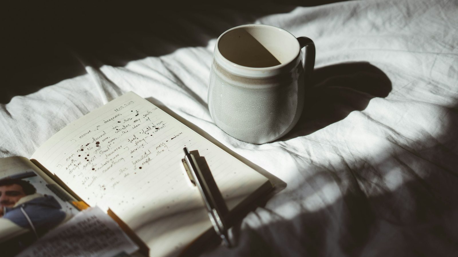 A mug of coffee and an open notebook with a pen on a bed in morning light