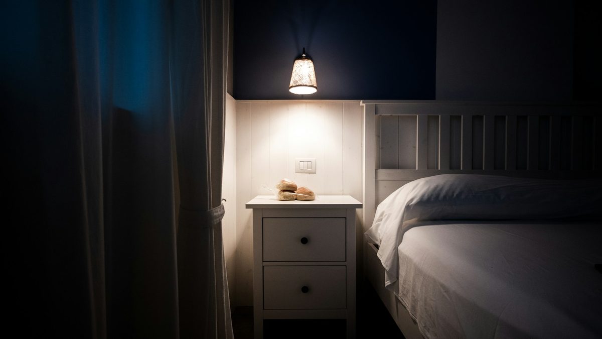 A single bedside lamp lighting an empty bed in a dark bedroom