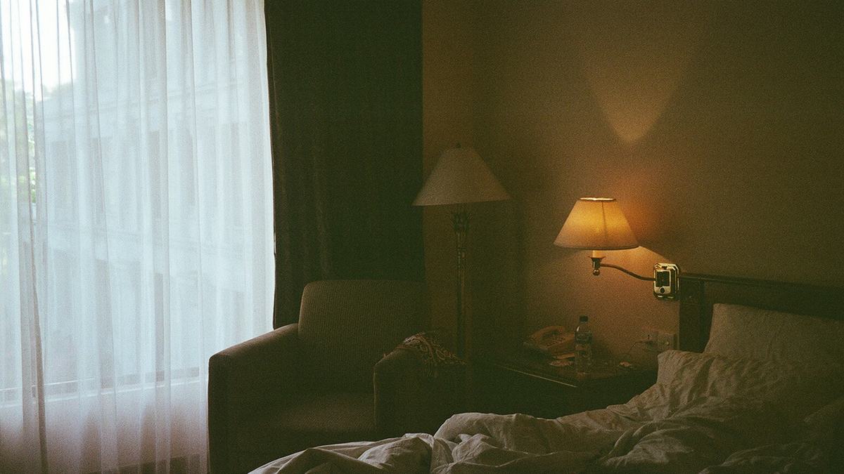 A dimly lit bedroom with a bed, an armchair and a warm bedside lamp