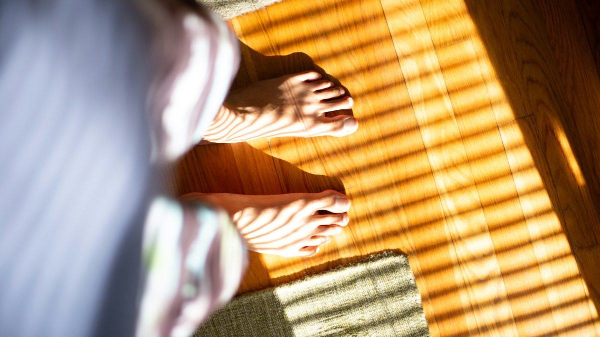 Bare feet standing on a sunlit wooden floor beside the bed