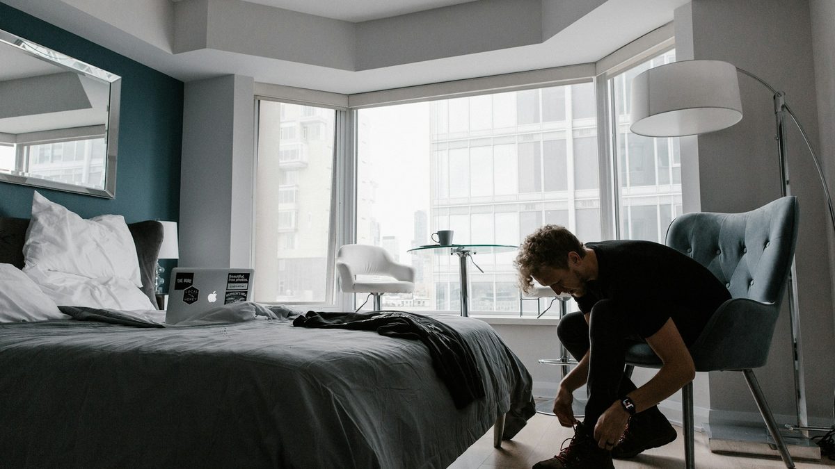 A man sitting on the edge of his bed tying his shoes in a bright morning bedroom
